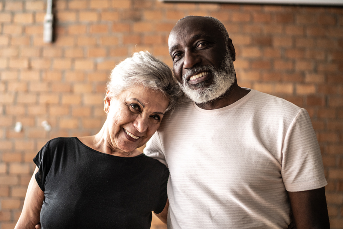 an elderly couple poses in front of a brick wall and smiles